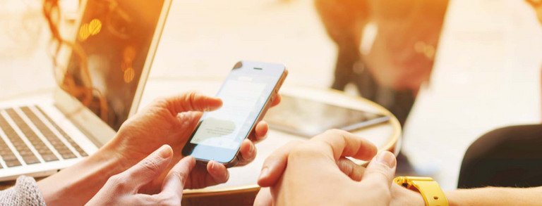 Zwei Hände halten Mobiltelefon, im Hintergrund steht ein Laptop
