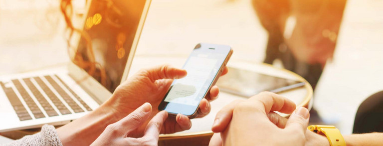 Zwei Hände halten Mobiltelefon, im Hintergrund steht ein Laptop