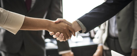 Handschlag Zwei Personen geben sich die Hand. Das Händeschütteln ist im Detail abgebildet, im Hintergrund stehen zwei Menschen im Anzug.