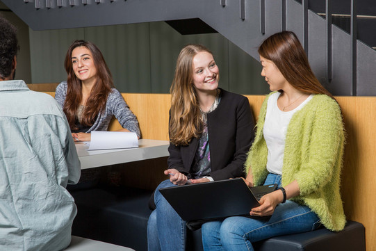 Zwei Studierende unterhalten sich und sitzen gemeinsam auf einer Bank mit einem Laptop.