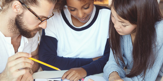 Drei Studierende haben ihre Köpfe über Unterlagen zusammengesteckt.