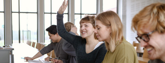Studierende im Seminar Fünf Studierende sitzen in einem Seminarraum an Tischen. Eine Studentin hebt ihre Hand.