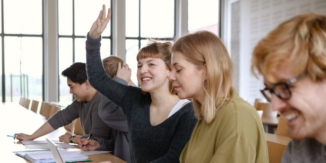 Fünf Studierende sitzen in einem Seminarraum an Tischen. Eine Studentin hebt ihre Hand.