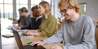 Studierende im Seminarraum sitzen in einer Tischreihe vor ihren Laptops. 